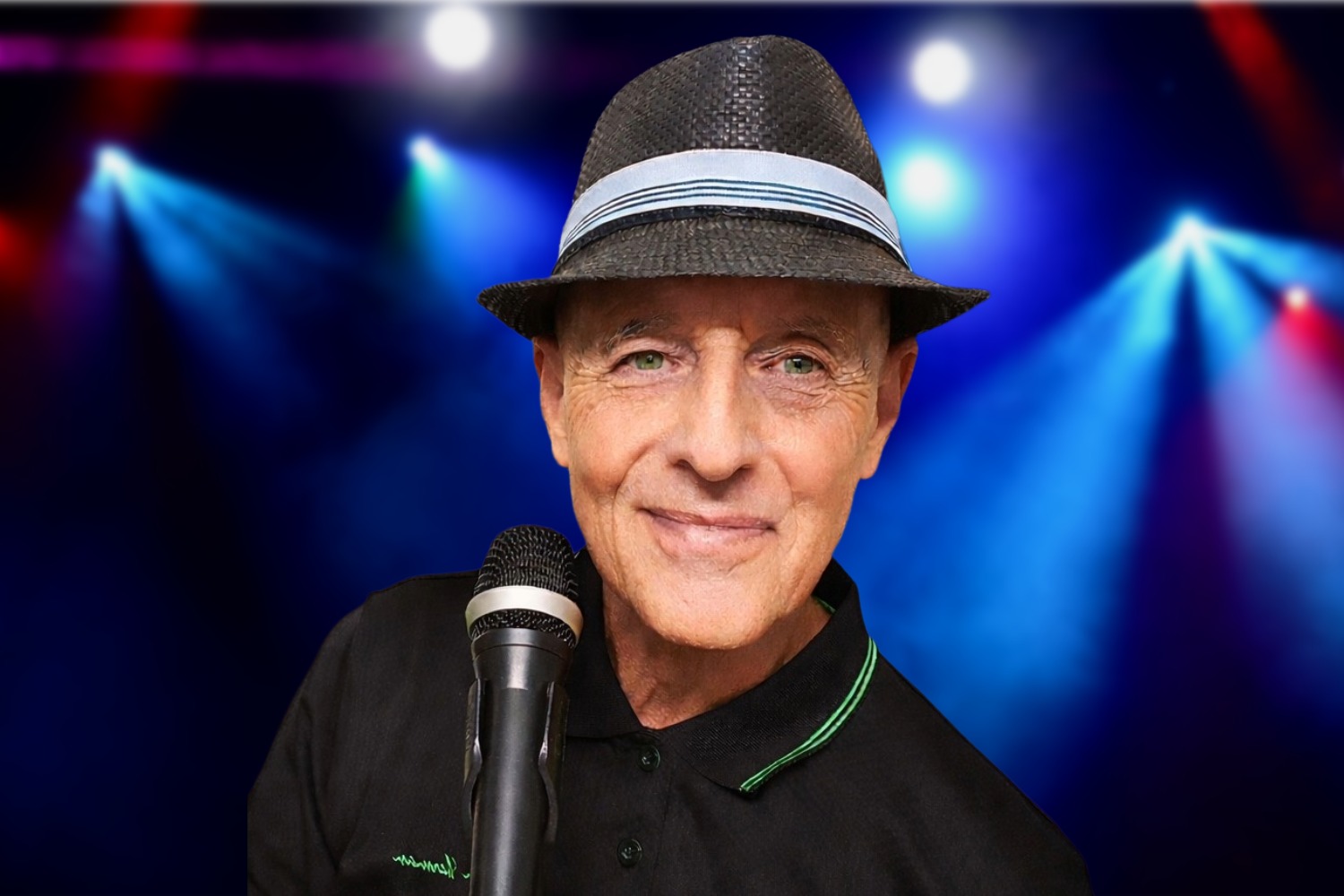 A close-up of a smiling man with green eyes wearing a dark woven fedora hat with a striped band. He stands behind a microphone against a digital background of vibrant blue and purple stage spotlights